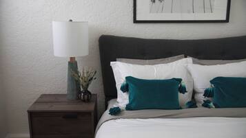 Een keurig opgemaakt bed met witte en teal kussens, een donker hoofdbord en een nachtkastje met een lamp en een vaas met gedroogde planten.