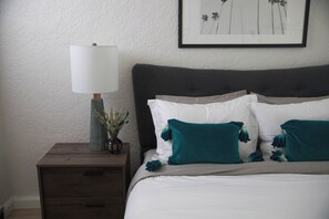 A neatly made bed with white and teal pillows, a dark headboard, and a bedside table with a lamp and a vase of dried flowers. - Cozy studio in fantastic Los Angeles with refreshing AC (Los Angeles)