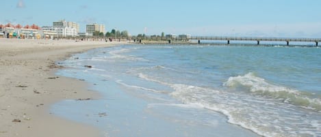 A beach with a pier extending into the water, buildings in the background, and a clear sky.