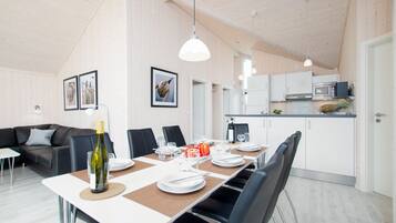 A modern dining area with a long table set for six, a sofa, and a kitchen in the background.
