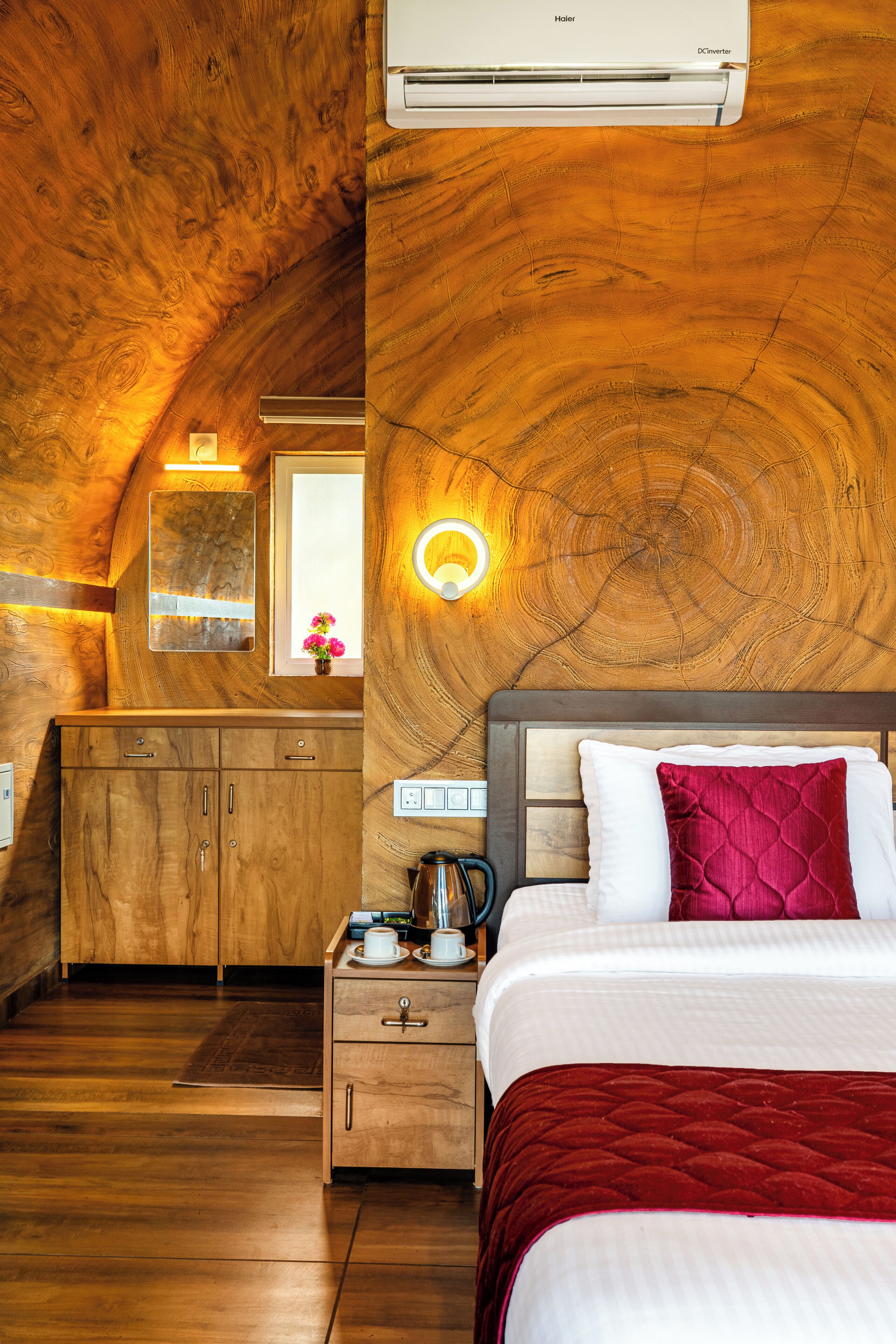 Ein Schlafzimmer mit Holzbalkendecke, einem Bett mit weißer und roter Bettwäsche, einem Nachttisch mit Lampe und einem Fenster mit Blumen.