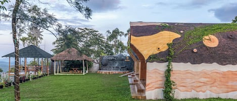 A grassy area with a thatched-roof structure, a wooden building with a unique design, and a stone sculpture.
