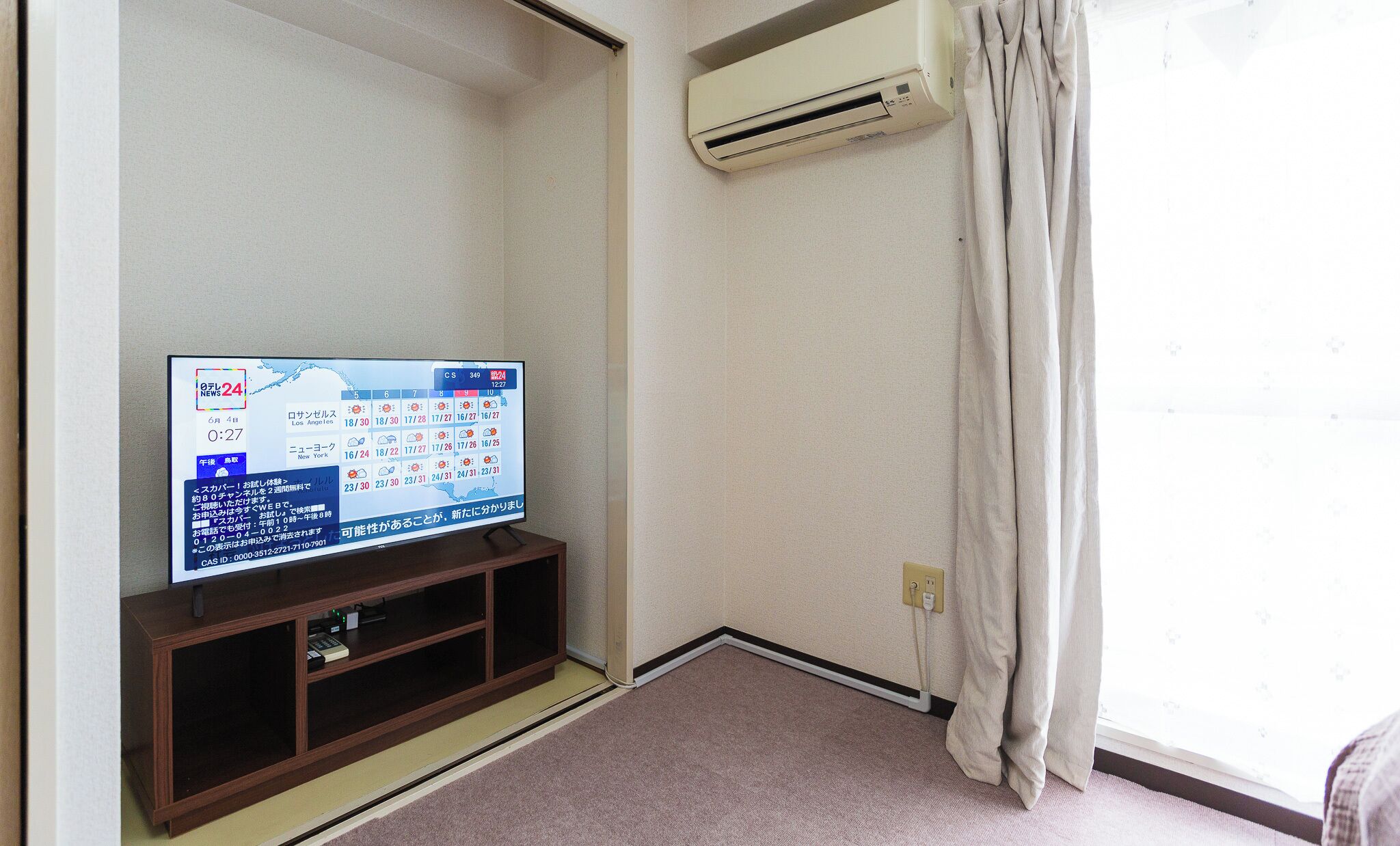 Un televisor sobre un soporte de madera con pantalla digital, un equipo de aire acondicionado montado en la pared y una ventana con cortinas.