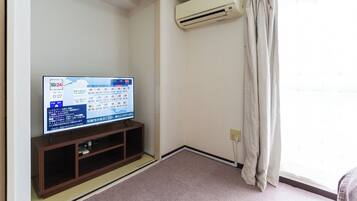 Un televisor sobre un soporte de madera con pantalla digital, un equipo de aire acondicionado montado en la pared y una ventana con cortinas.