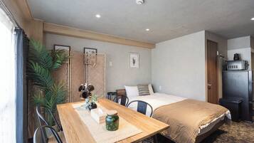 Habitación de hotel compacta con cama, escritorio, silla, una mesita, planta en maceta y kitchenette.