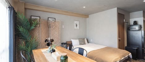 A compact hotel room with a bed, a desk, a chair, a small table, a potted plant, and a kitchenette.