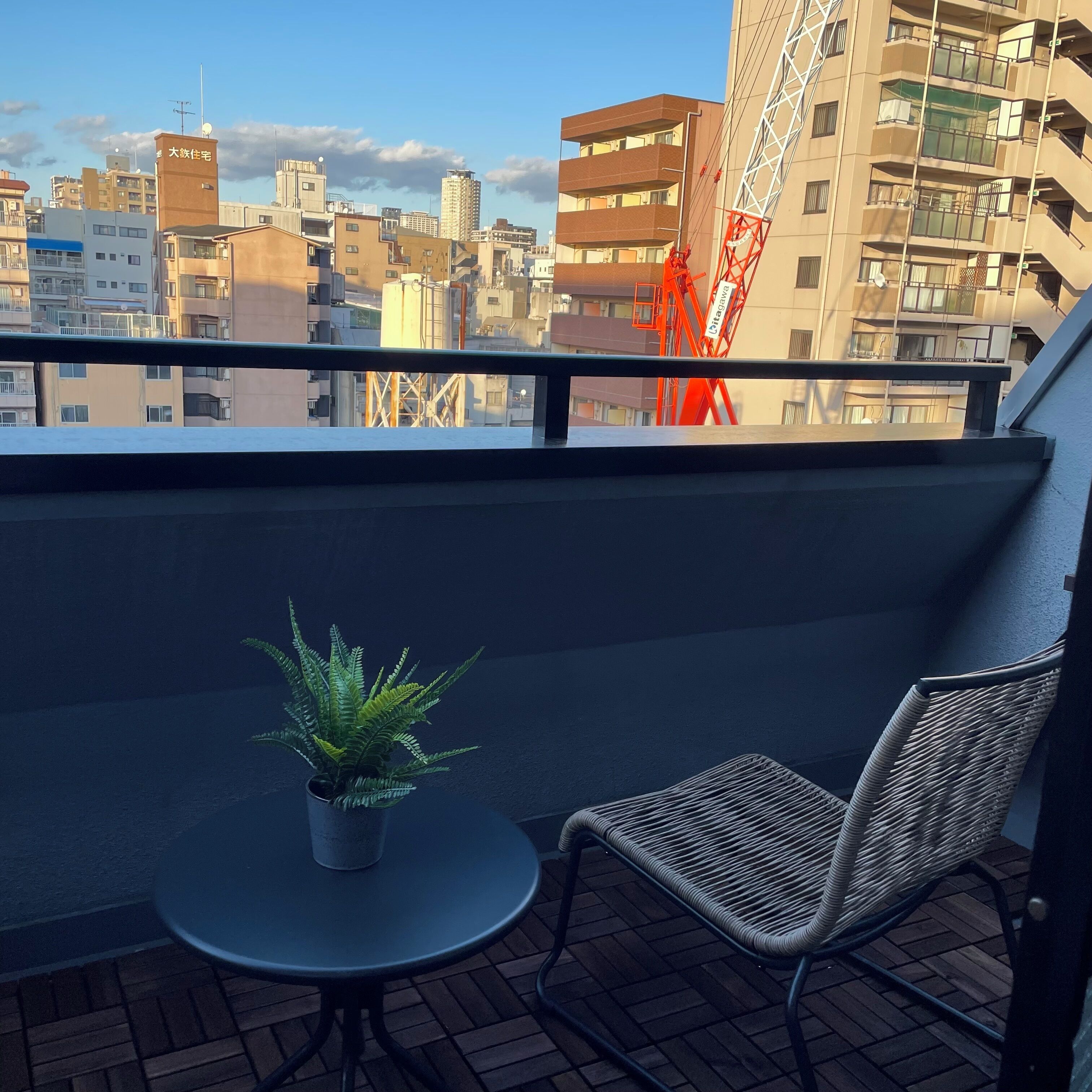 A balcony with a potted plant and a wicker chair, overlooking a cityscape with buildings and a crane.