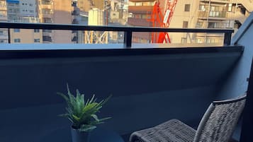 A balcony with a potted plant and a wicker chair, overlooking a cityscape with buildings and a crane.