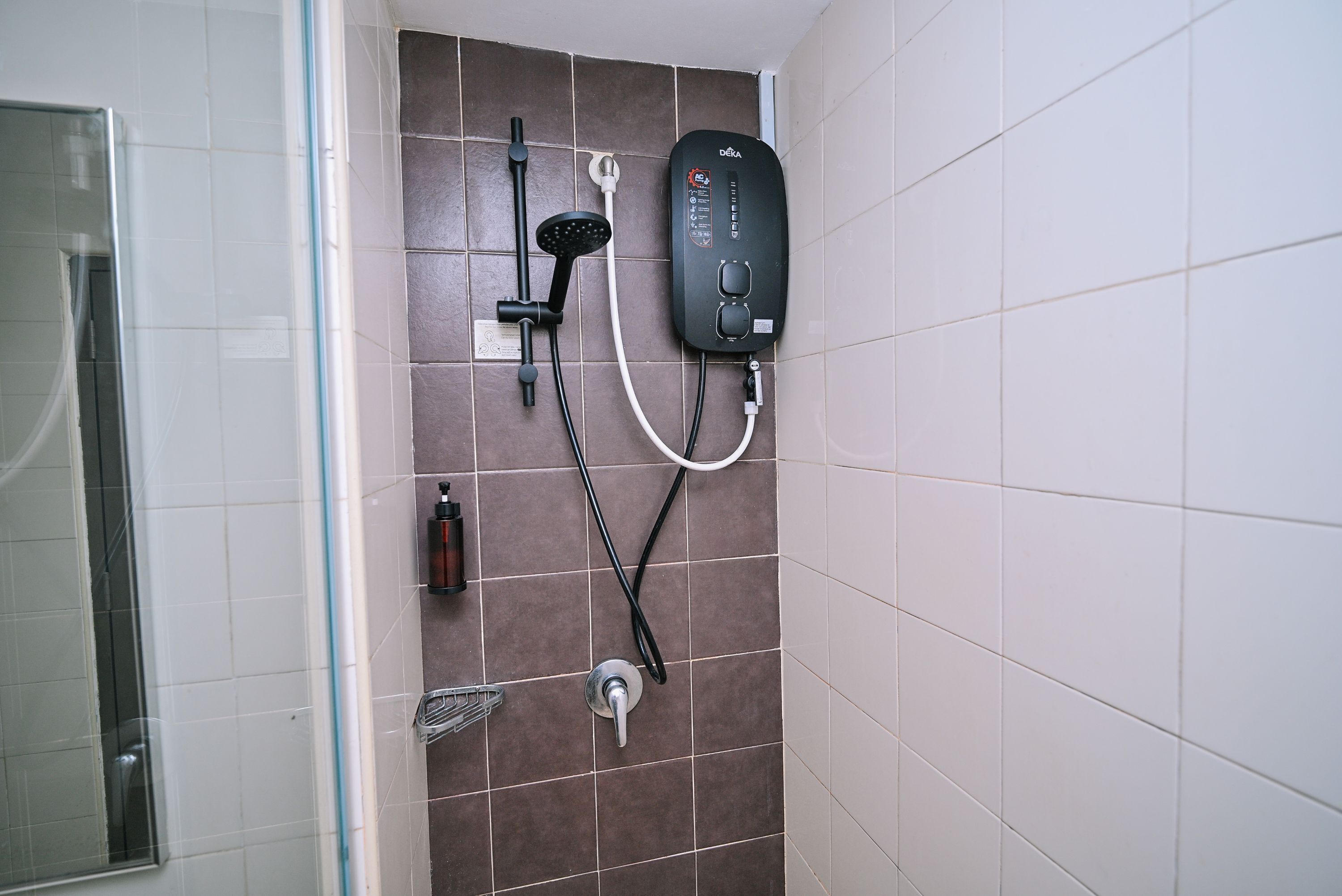 A bathroom with a shower, a handheld showerhead, a wall-mounted faucet, and a soap dispenser.
