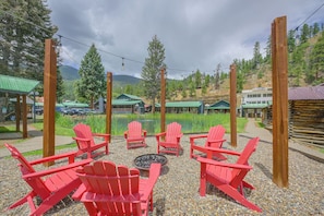 A seating area with red chairs and a fire pit, surrounded by a gravel path and greenery, with buildings and a lake in the background. - Walk to Main St: Red River Lodge w/ Ski Views! (Red River)