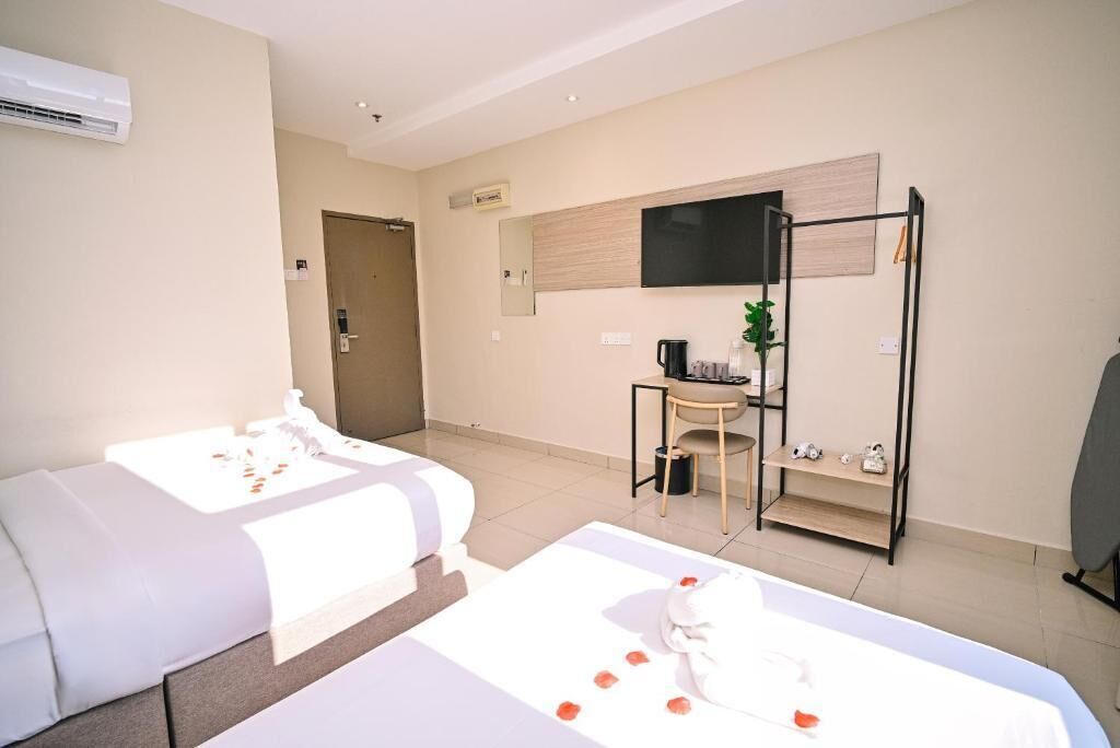 A hotel room with two beds, a desk, a chair, a TV, and a small shelf with items.