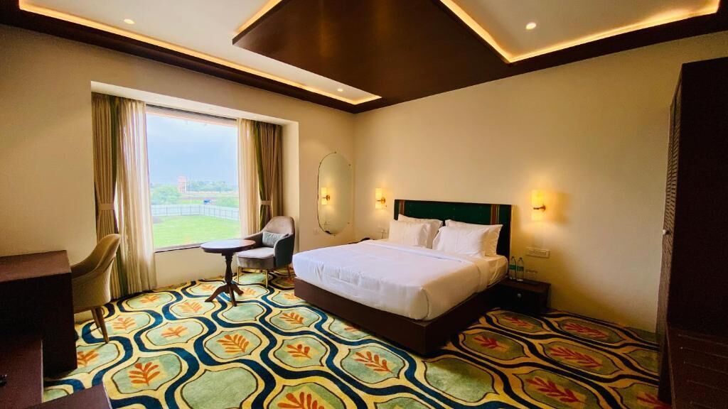 A hotel room with a bed, a desk, a chair, a large window, and a patterned carpet.