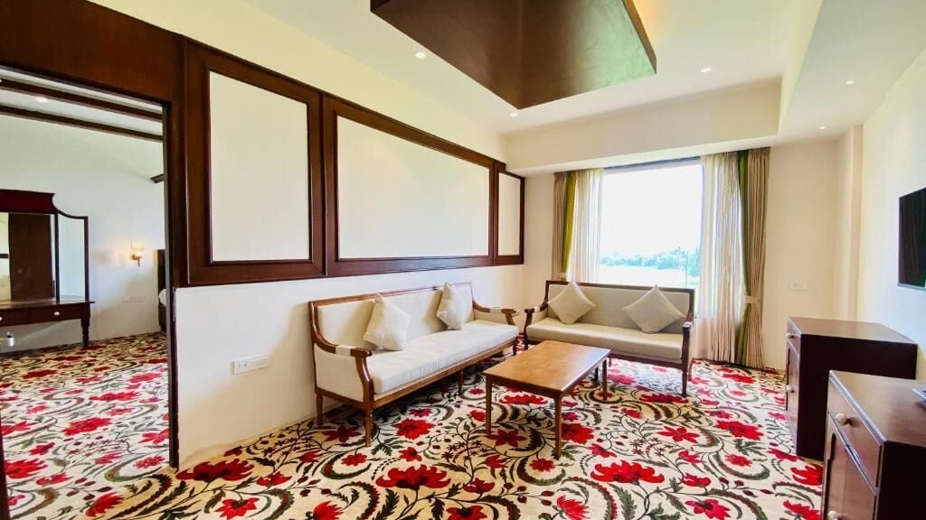Quarto de hotel com sofá, mesa de centro, televisão e uma janela grande com cortinas.