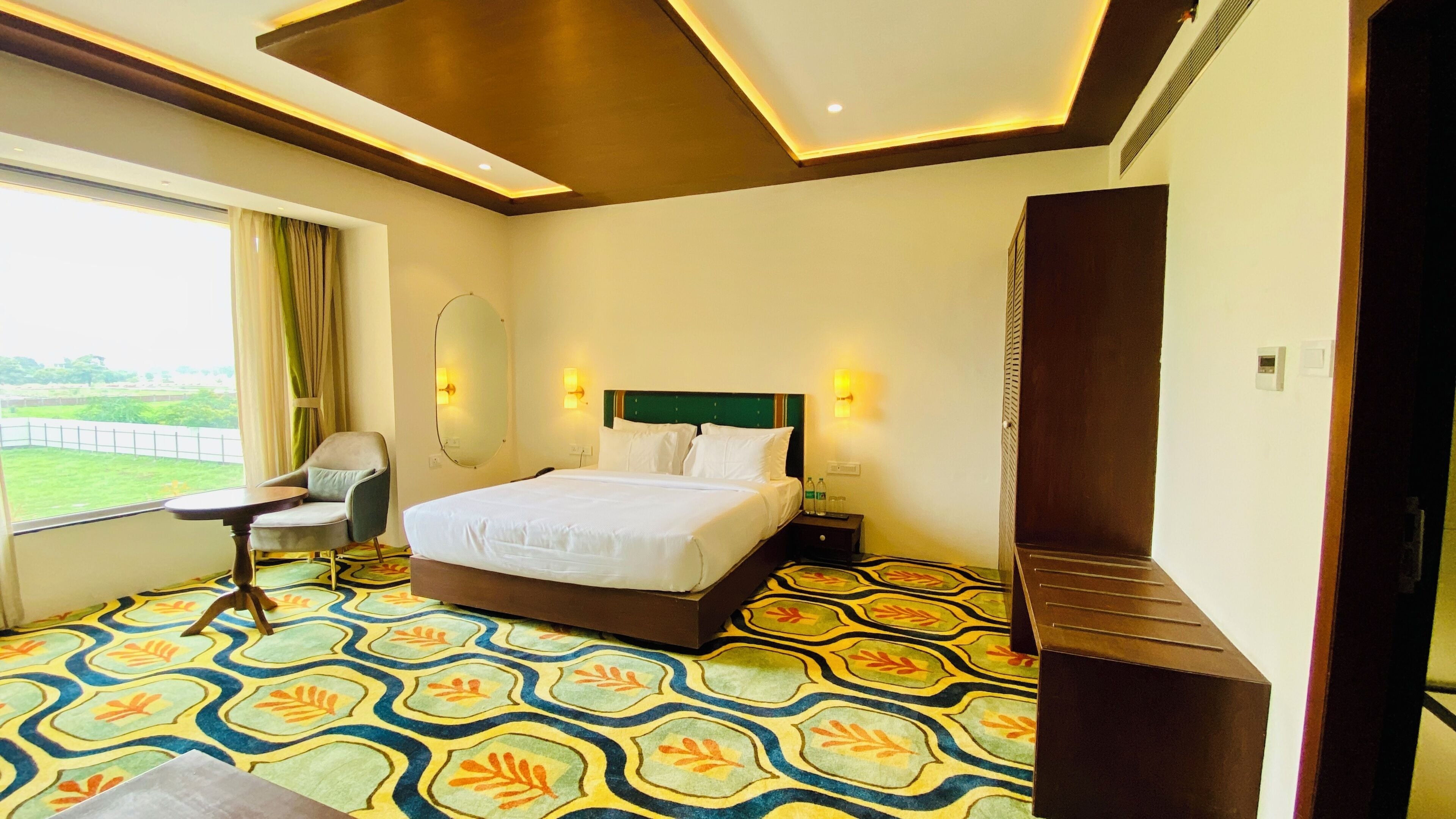 A hotel room with a large bed, a chair, a desk, a mirror, and a view of a green landscape.