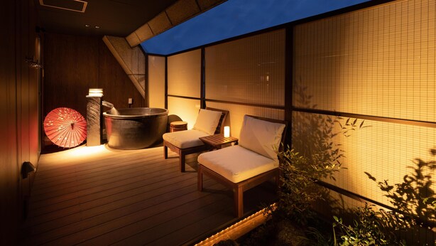 Terraza de madera con un jacuzzi, dos sillones acolchados, un paraguas rojo y una habitación tradicional japonesa con esteras de tatami.