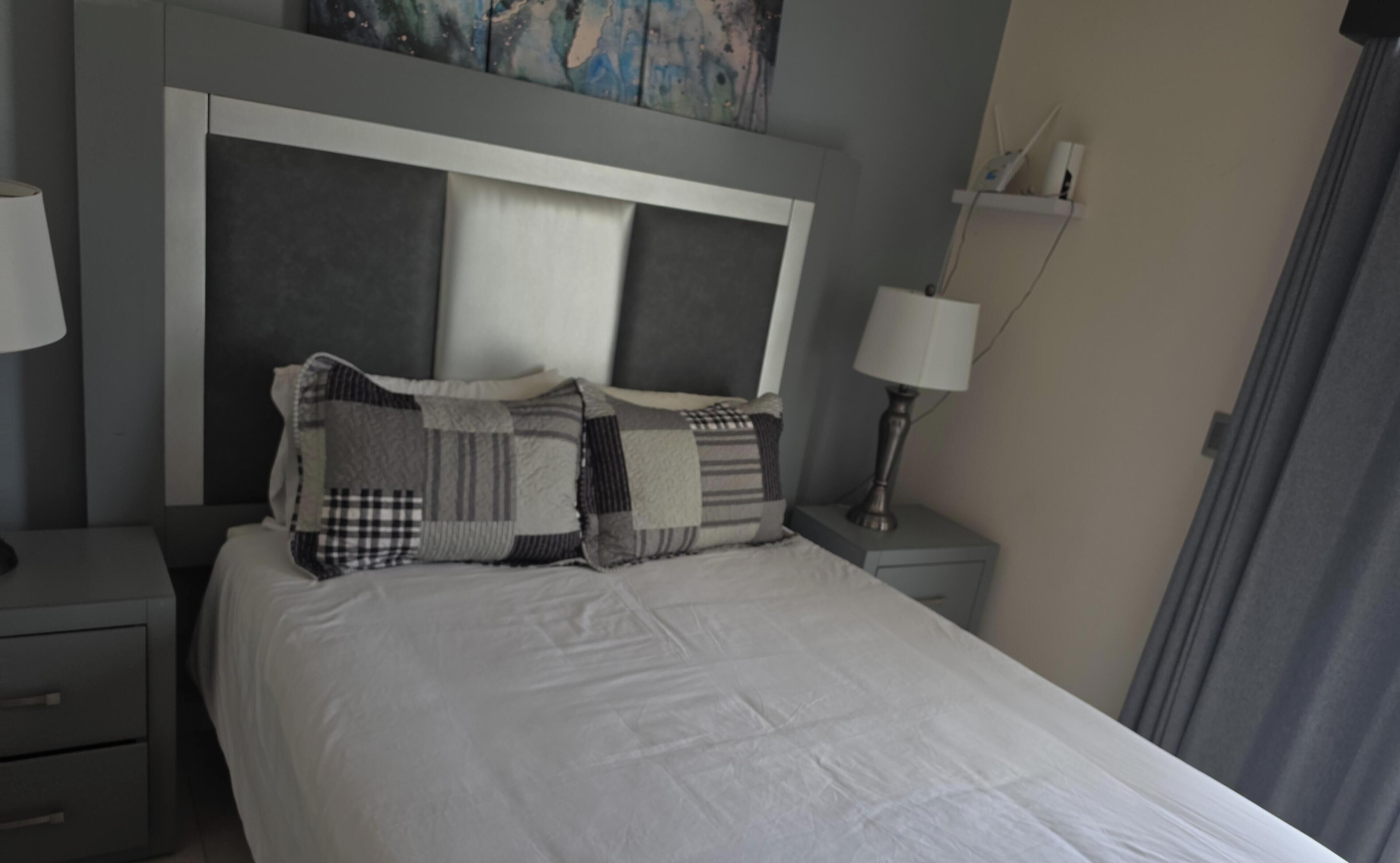 A neatly made bed with a white duvet, a grey headboard, and multiple pillows. There are two bedside tables, each with a lamp and a small shelf.