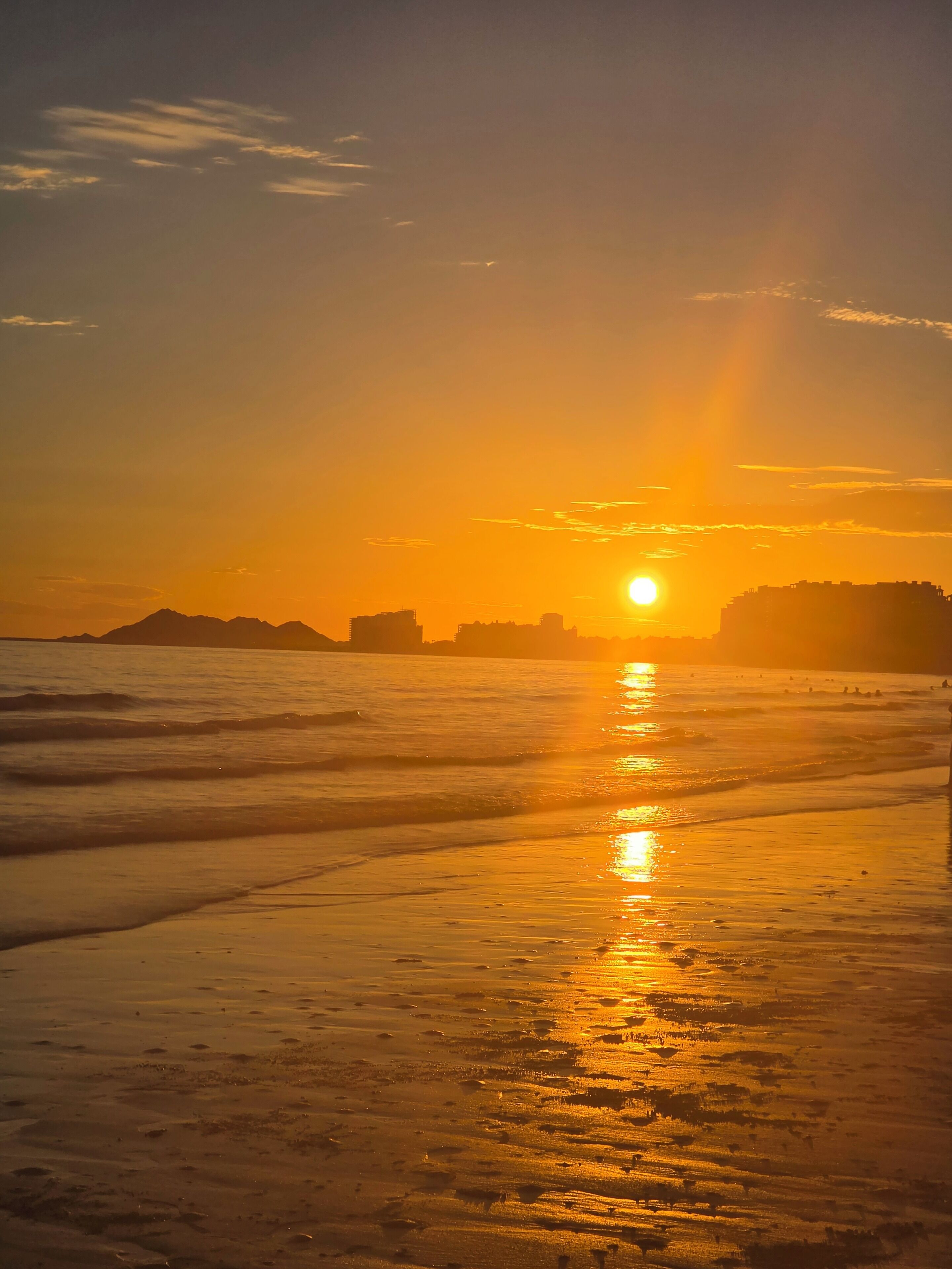 A sunset over a beach with waves and a distant city skyline.