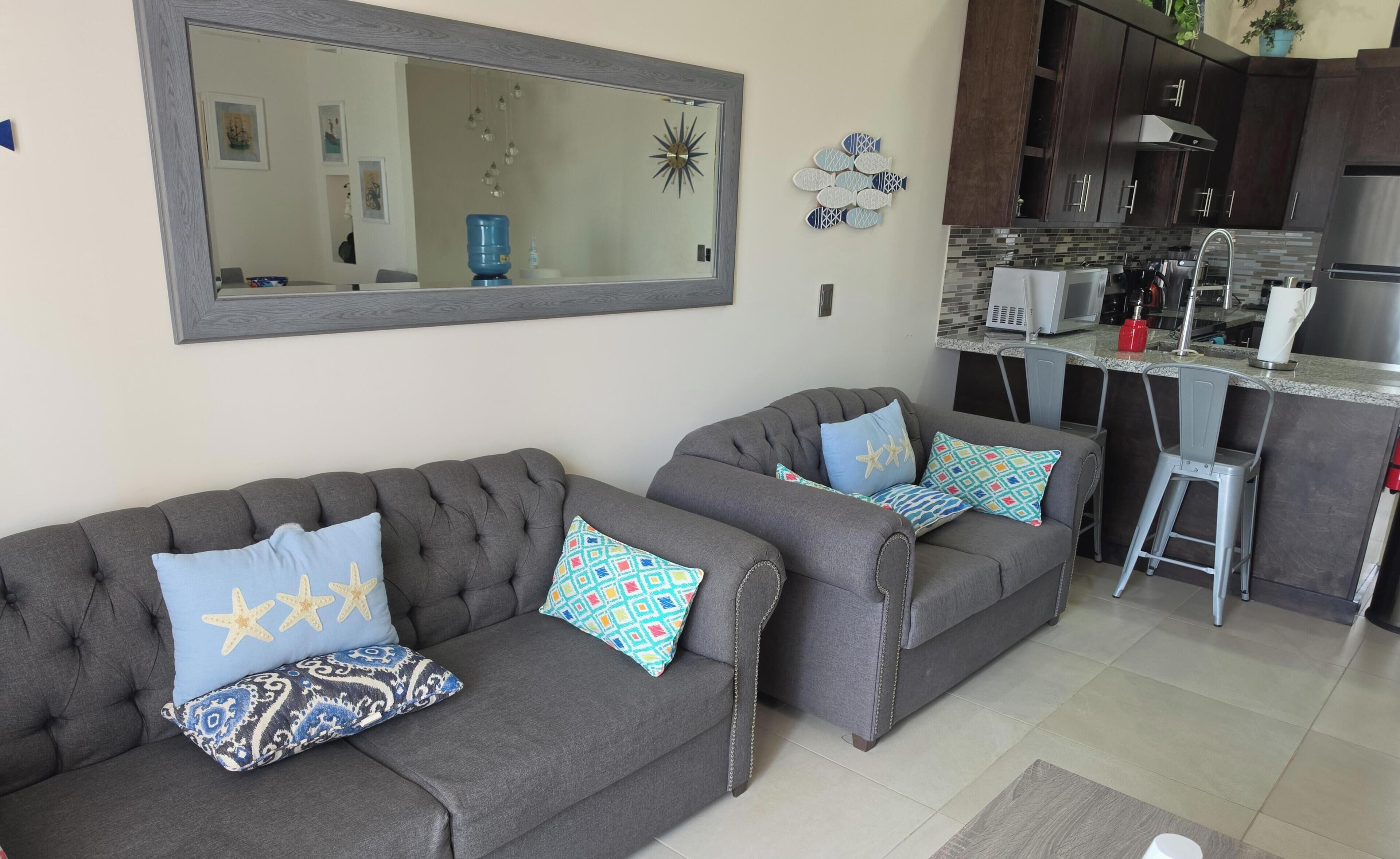 A living room with two grey sofas, each with patterned cushions, a large mirror above, and a kitchen area with modern appliances and a tiled backsplash.