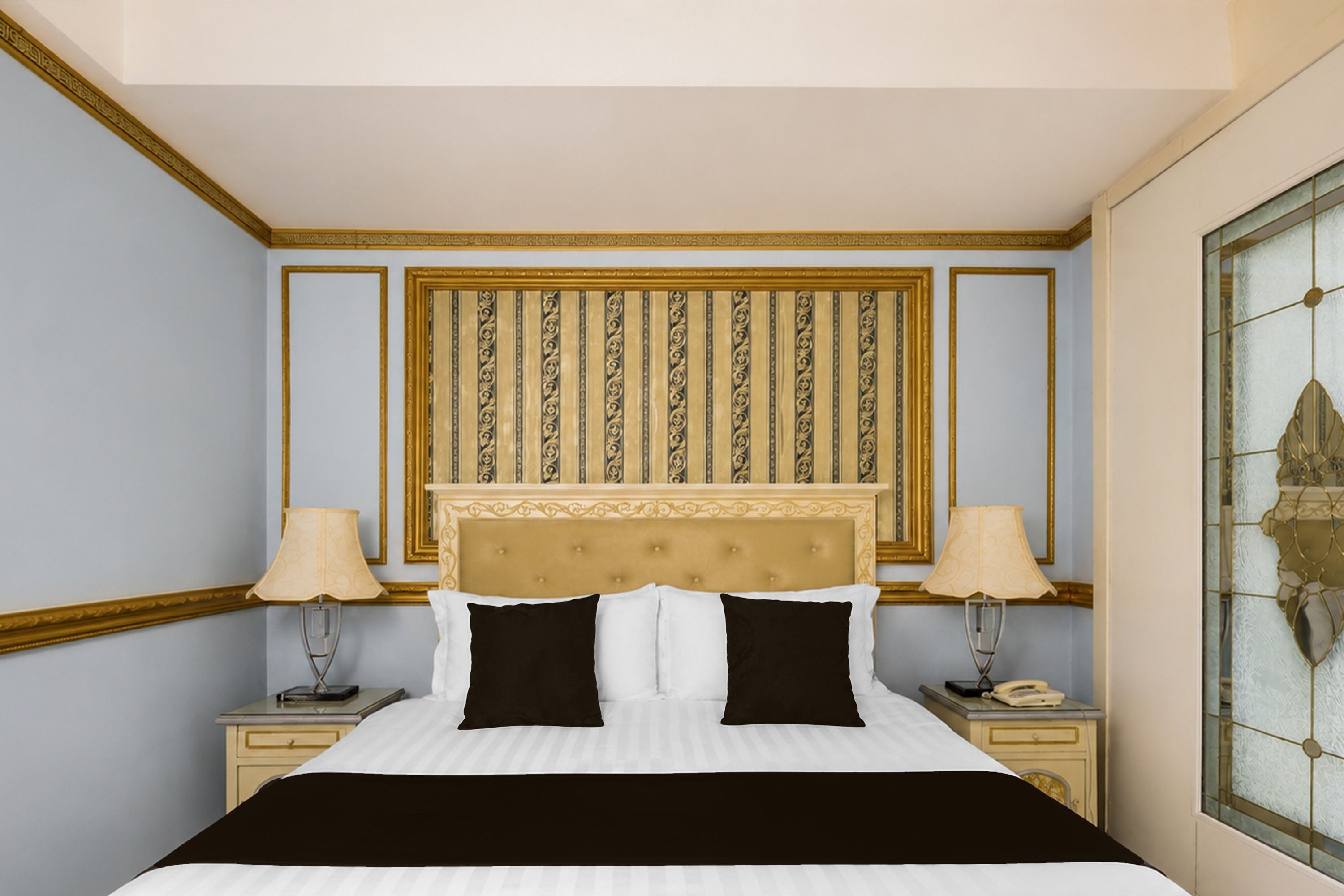 A hotel room with a large bed, two bedside tables with lamps, a decorative headboard, and a window with a geometric pattern.
