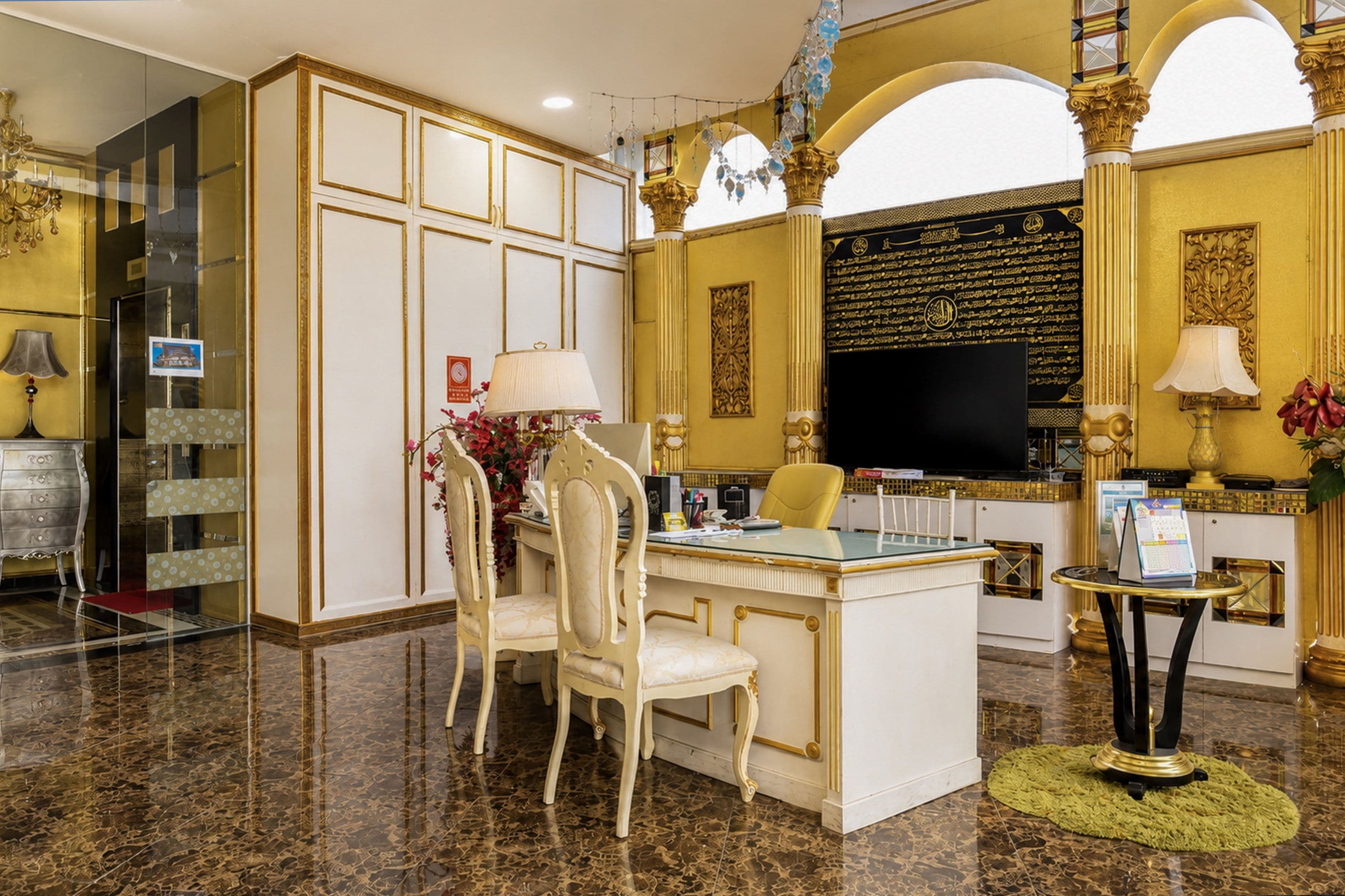 A luxurious hotel lobby with marble flooring, a central desk with chairs, a television, and decorative elements.