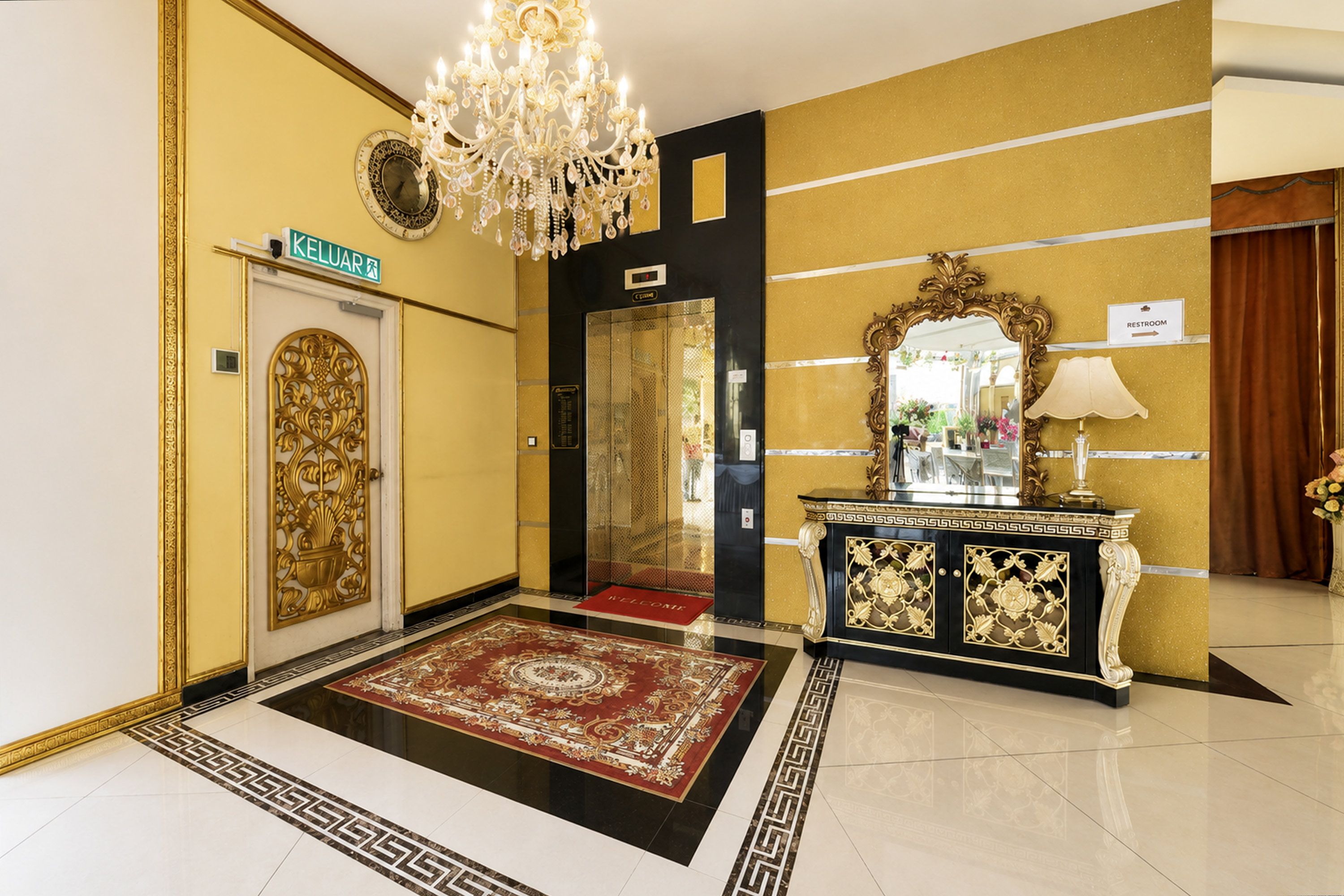 Um hall de hotel com um candelabro, um tapete vermelho, um espelho com moldura dourada e uma placa indicando a localização.