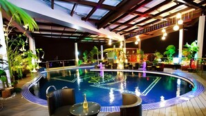 A swimming pool with a sunken center, surrounded by lounge chairs and tables, under a covered area with wooden beams and hanging lights.