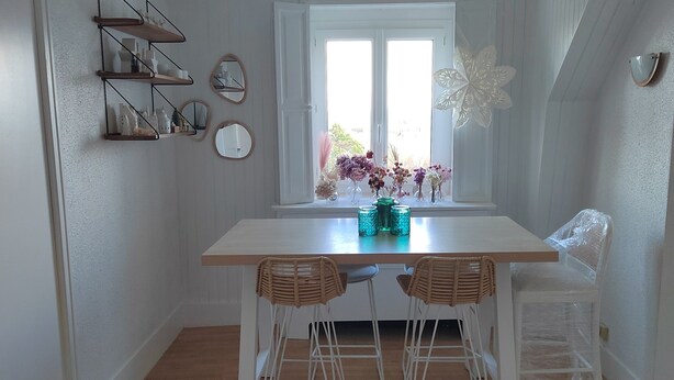 A dining area with a white table, two wicker chairs, a window with a view, and wall-mounted shelves.