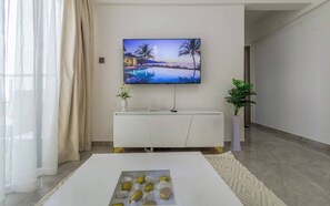A modern living room with a flat-screen TV mounted on the wall, a white entertainment unit below, a coffee table with decorative stones, and a potted plant to the side. - Cozy Nest 1BR Kilimani (Nairobi)