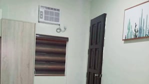 A room with a wardrobe, a desk with a chair, a window with blinds, a door, and a wall-mounted air conditioning unit.