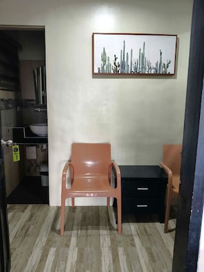 A room with a wooden floor, a plastic chair, a black cabinet, and a painting of cacti on the wall.