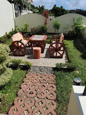 Un espace jardin agrémenté d’une table d’échecs et de chaises, entouré de verdure et bordé d’un chemin pavé.