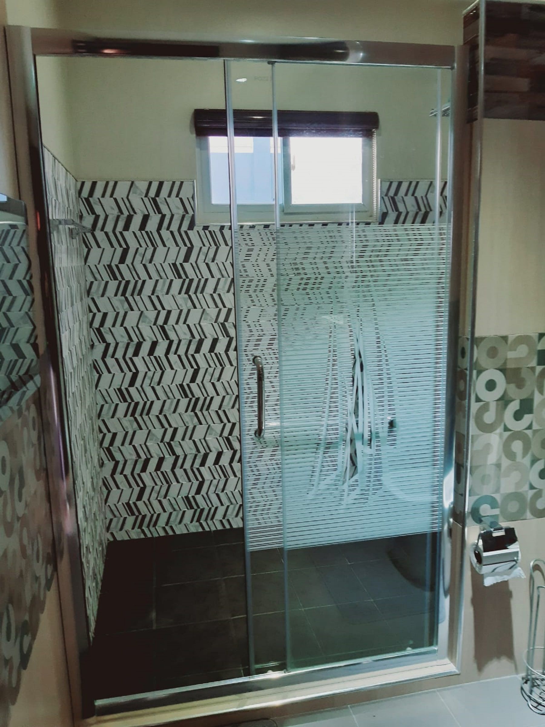 A shower enclosure with a glass door, patterned walls, and a window above.