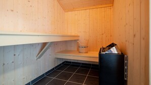 A small, enclosed space with wooden walls and a tiled floor, featuring a shelf, a trash bin, and a chair. - Grobenbrode House in der Nähe von Shore (Großenbrode)