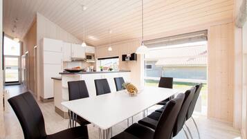 Eine moderne Küche mit weißen Schränken, ein Essbereich mit einem weißen Tisch und schwarzen Stühlen sowie ein Blick auf ein Haus durch ein großes Fenster.