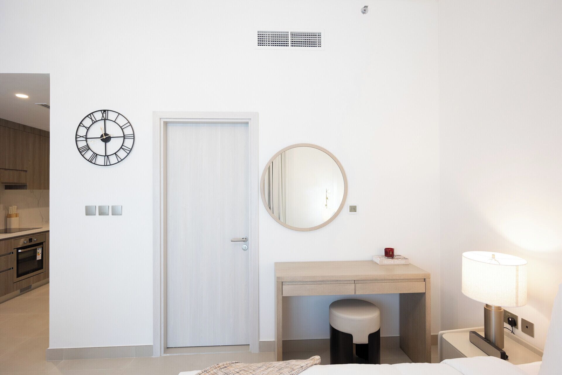 Une chambre d’hôtel comprenant un lit, une table de chevet avec une lampe, un miroir rond, un petit bureau avec une chaise et une horloge murale.