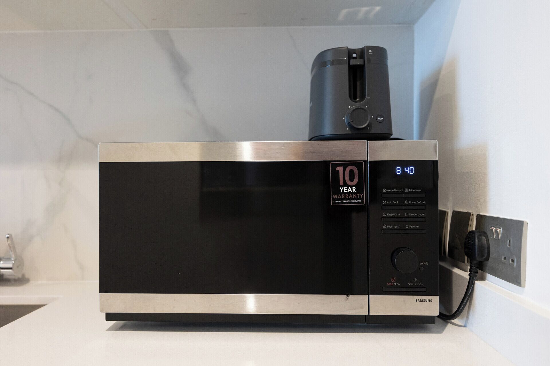 A stainless steel microwave oven with a digital display, a 10-year warranty badge, and a power cord plugged in.