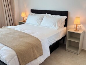 Un lit bien fait, avec du linge de lit blanc et un plaid beige, flanqué de deux tables de chevet avec des lampes, dans une pièce au design simple et épuré.