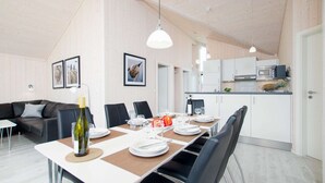 Uma zona de refeições moderna com uma mesa comprida posta para quatro pessoas, rodeada por cadeiras, e uma cozinha ao fundo.