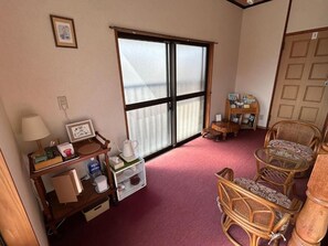 Una habitación con un ventanal grande, una silla de madera, una mesita y una estantería con diversos objetos.