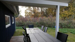 Um pátio externo com mesa e cadeiras de madeira, rodeado por um jardim com árvores e arbustos.