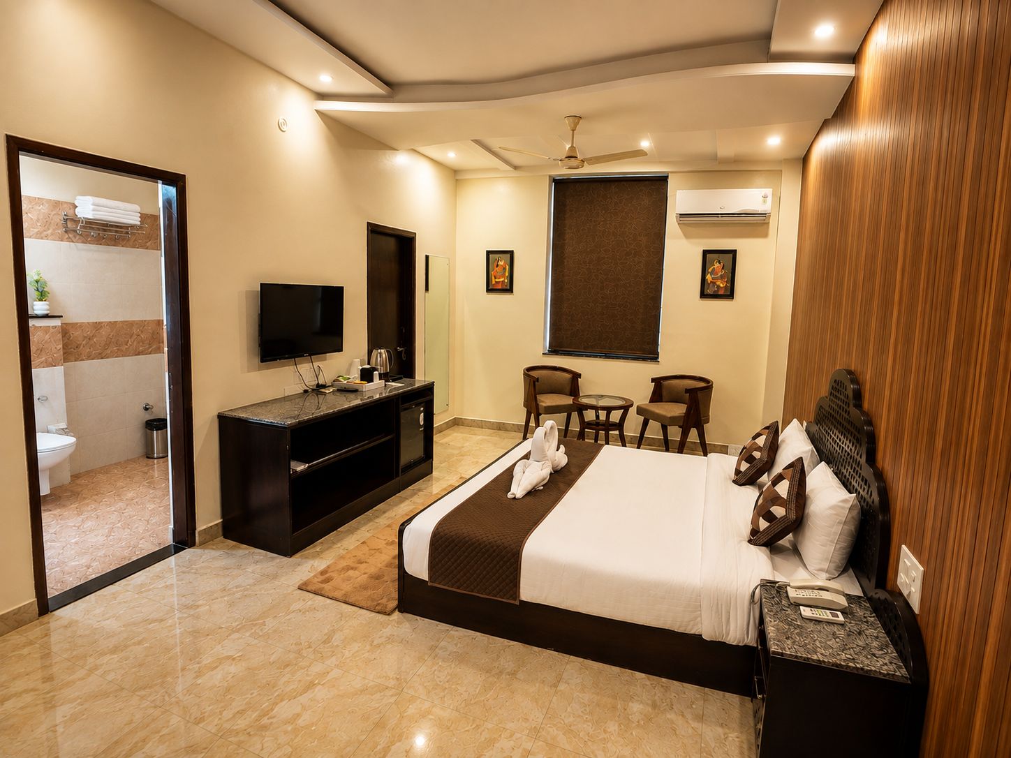 Habitación de hotel con cama, escritorio, televisión, una mesita, sillas y baño visible a través de una puerta abierta.