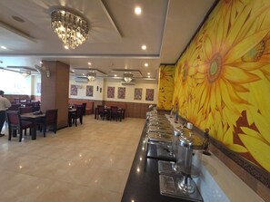 A restaurant with a long counter, multiple tables, and chairs. There are ceiling lights and a large sunflower mural on the wall. - Hote Elvora (Jaipur)