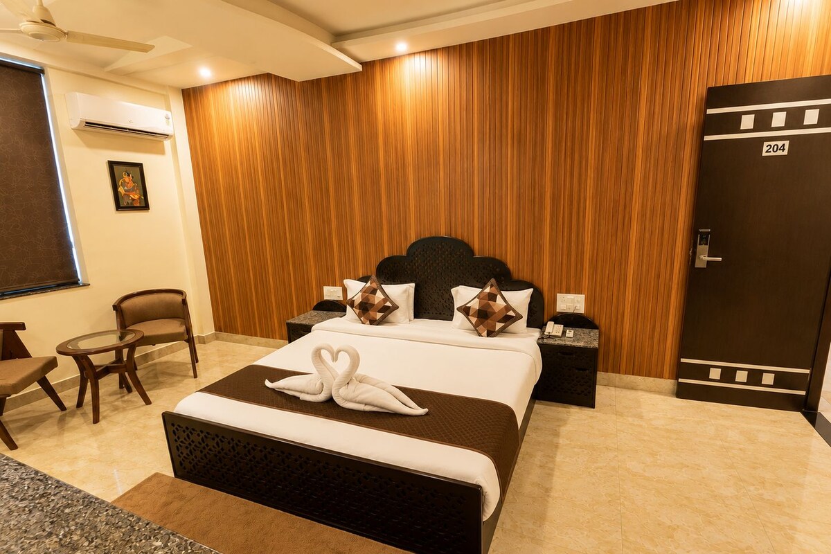 A hotel room with a bed, bedside tables, a chair, a small table, a wall-mounted air conditioner, and a door with a number.