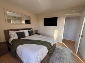A bedroom with a bed, a bedside table, a lamp, a flat-screen TV, a mirror, and a door.