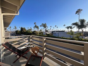 A balcony with two lounge chairs, a small table, and a view of palm trees and a street.