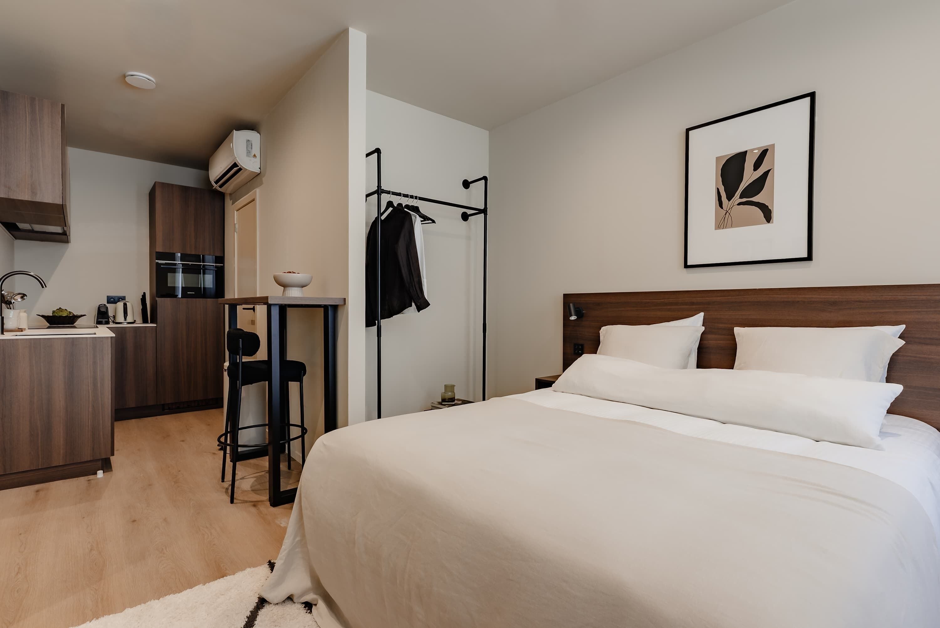Une chambre d’hôtel moderne comprenant un lit, une table de chevet, une chaise, un petit coin cuisine et une salle de bain.