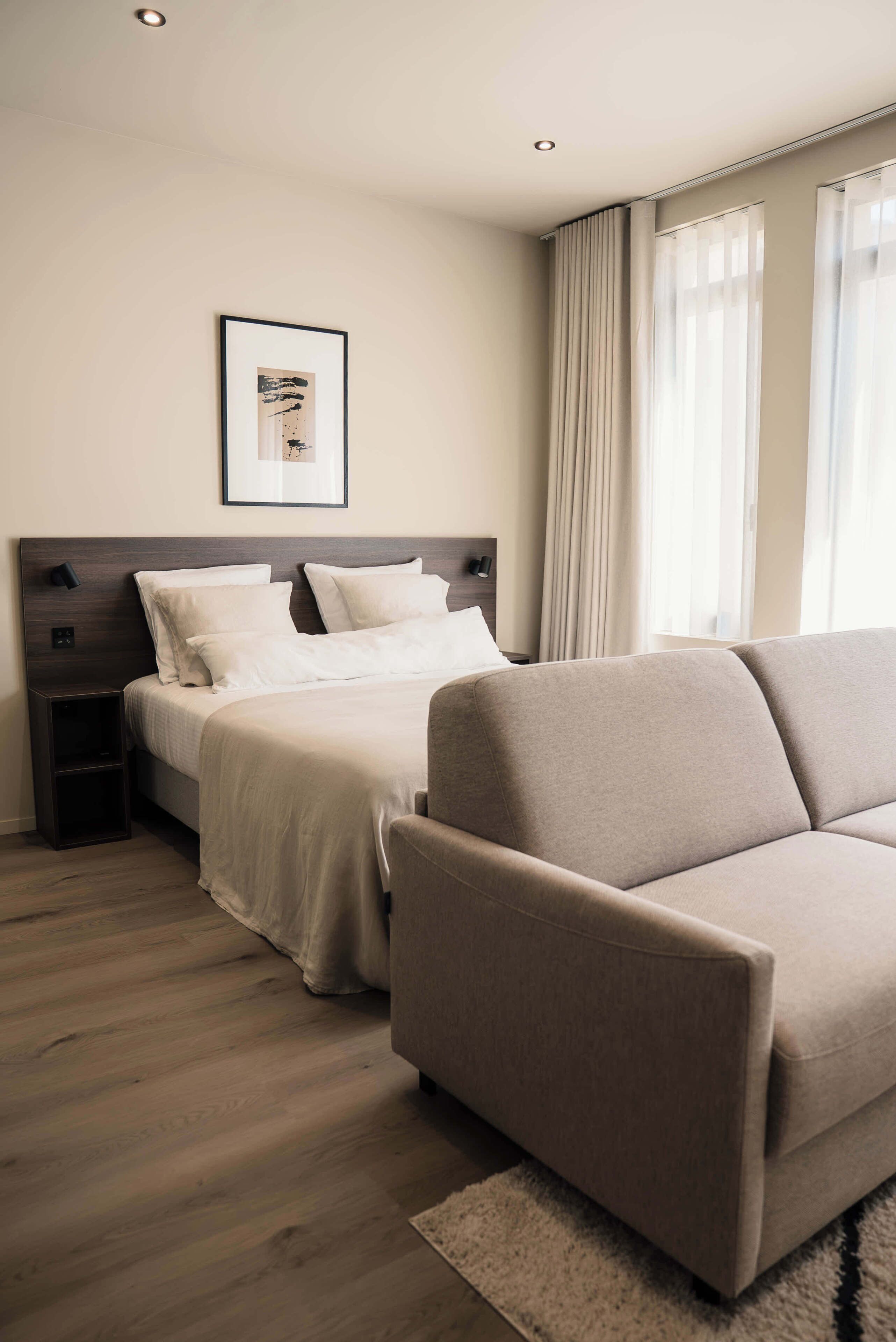 Une chambre d’hôtel avec un lit, un canapé, une table de chevet, un tableau encadré au mur et une fenêtre avec des rideaux.