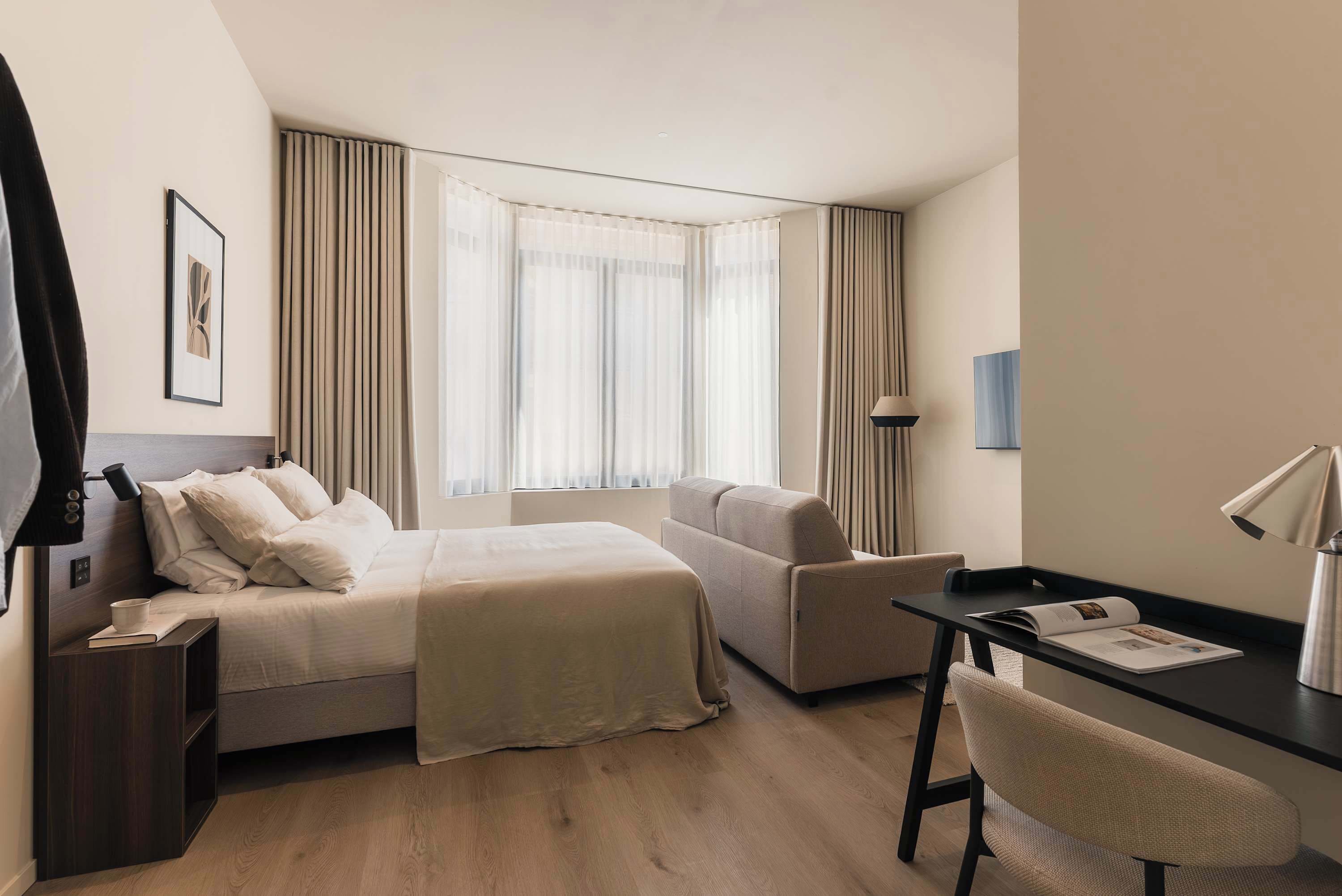 A modern hotel room with a bed, a desk, a chair, and a large window with curtains.