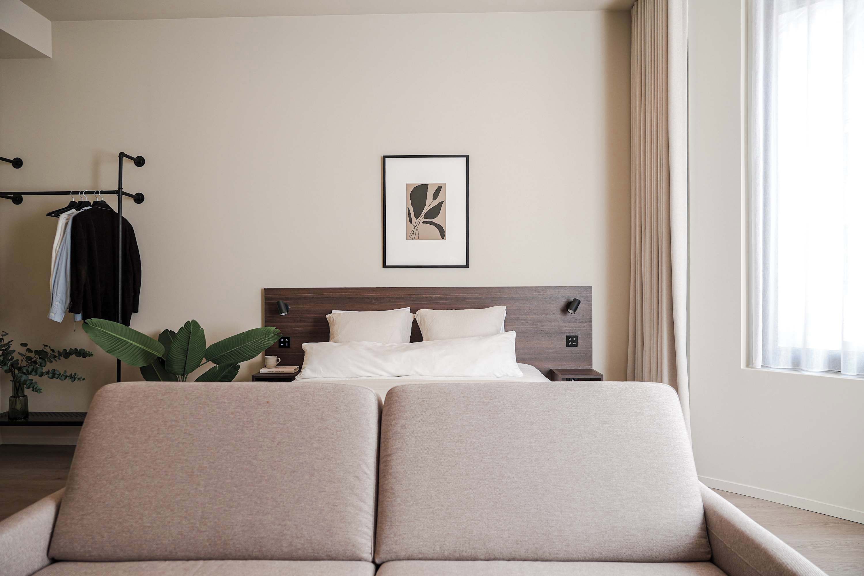 A modern bedroom with a bed, bedside tables, a wall-mounted artwork, a clothing rack, and a potted plant.