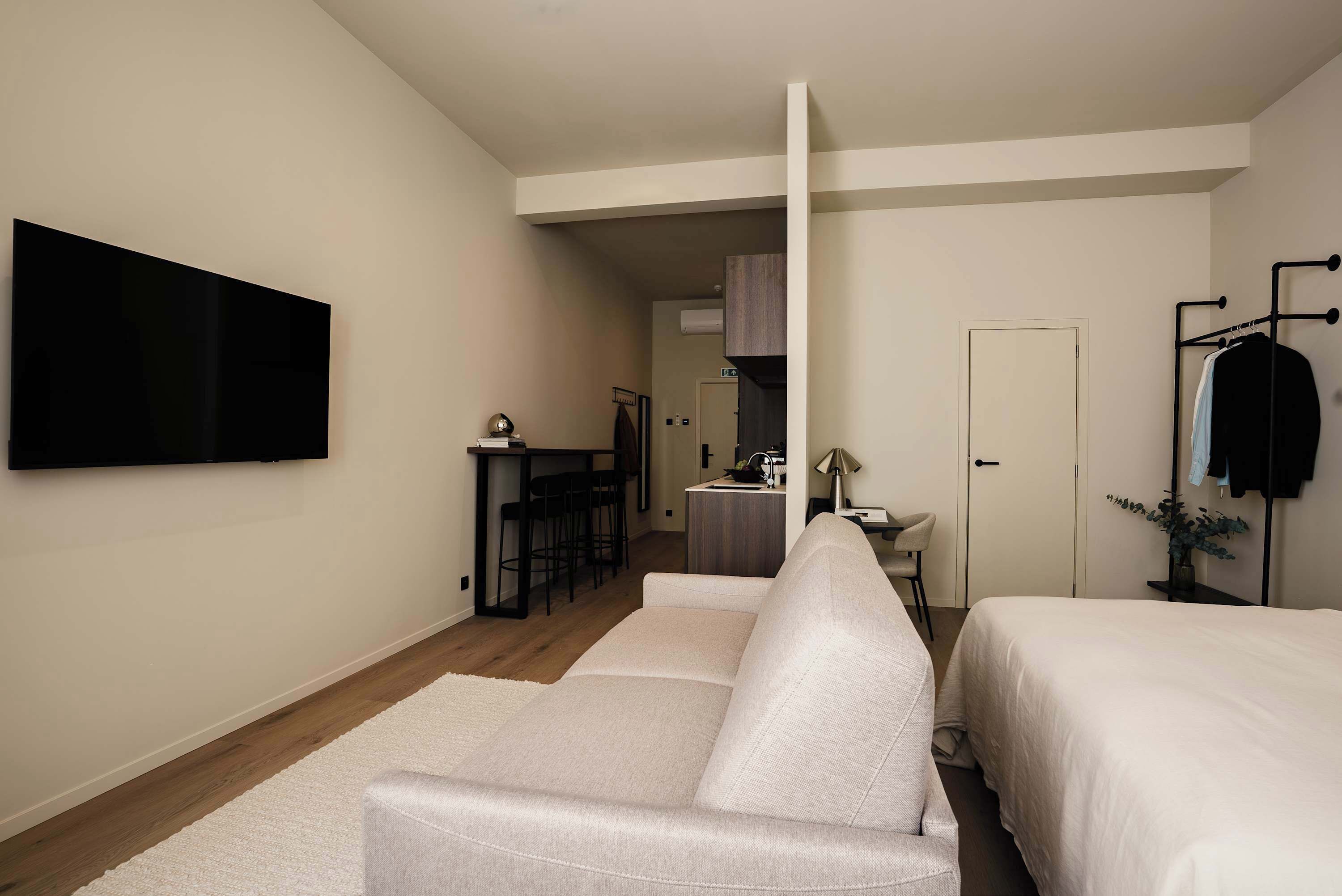 A modern hotel room with a flat-screen TV, a sofa, a bed, a desk, and a kitchenette.